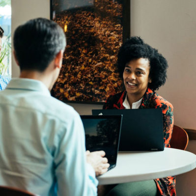 Two people sitting at a round table with laptops, engaged in conversation in a relaxed, cafe-like setting.